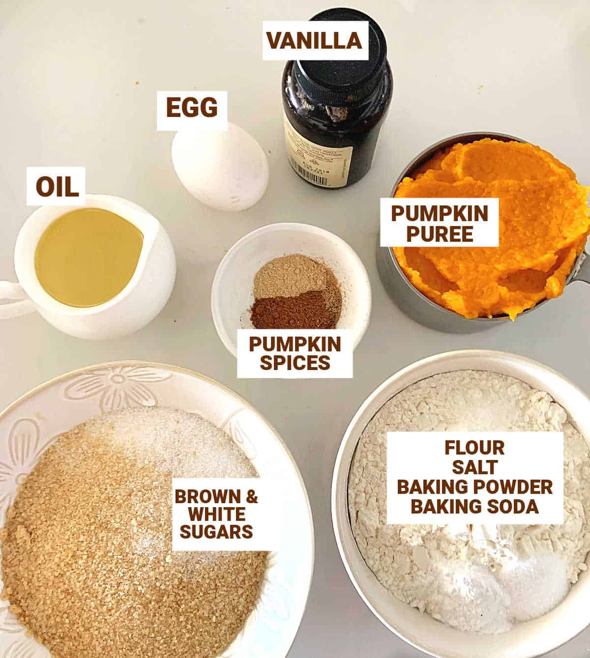 Whitish surface with bowls containing ingredients for pumpkin cookies including sugar, vanilla, oil, spices, flour, egg.