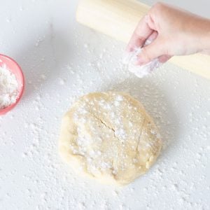 Hand dusting flour onto round of pie dough, rolling pin, pink bowl on a white surface