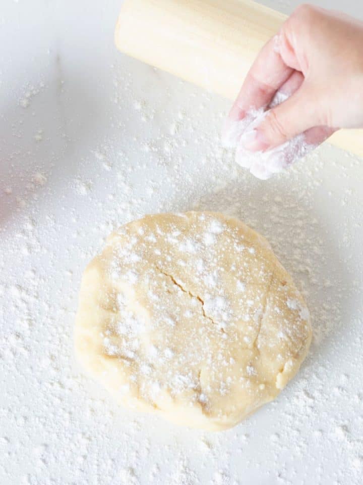 Hand dusting flour onto round of pie dough, rolling pin, pink bowl on a white surface