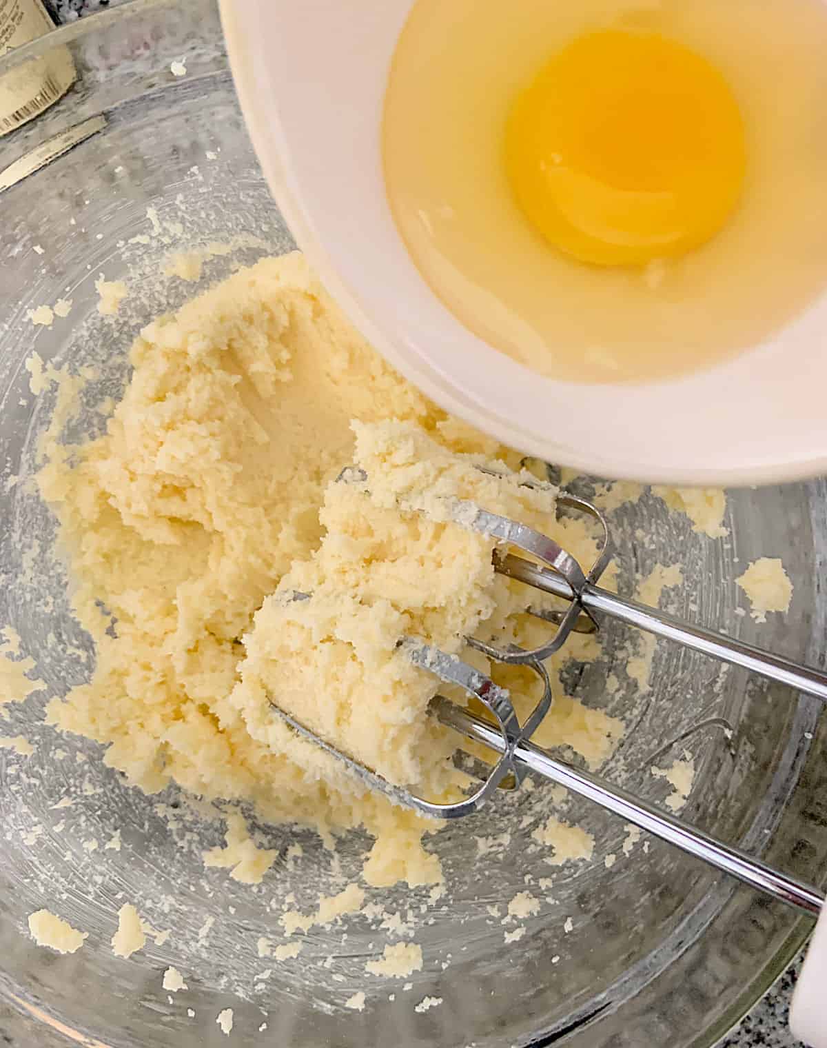 Adding egg from a white bowl to cake batter in glass bowl, beaters inside