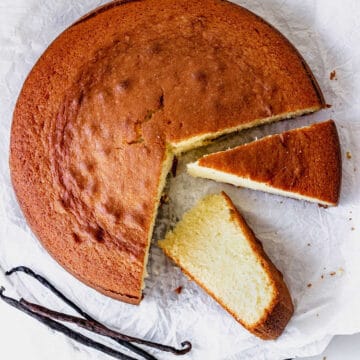 Top view of round plain vanilla cake on white paper, whole vanilla beans, two cut slices.