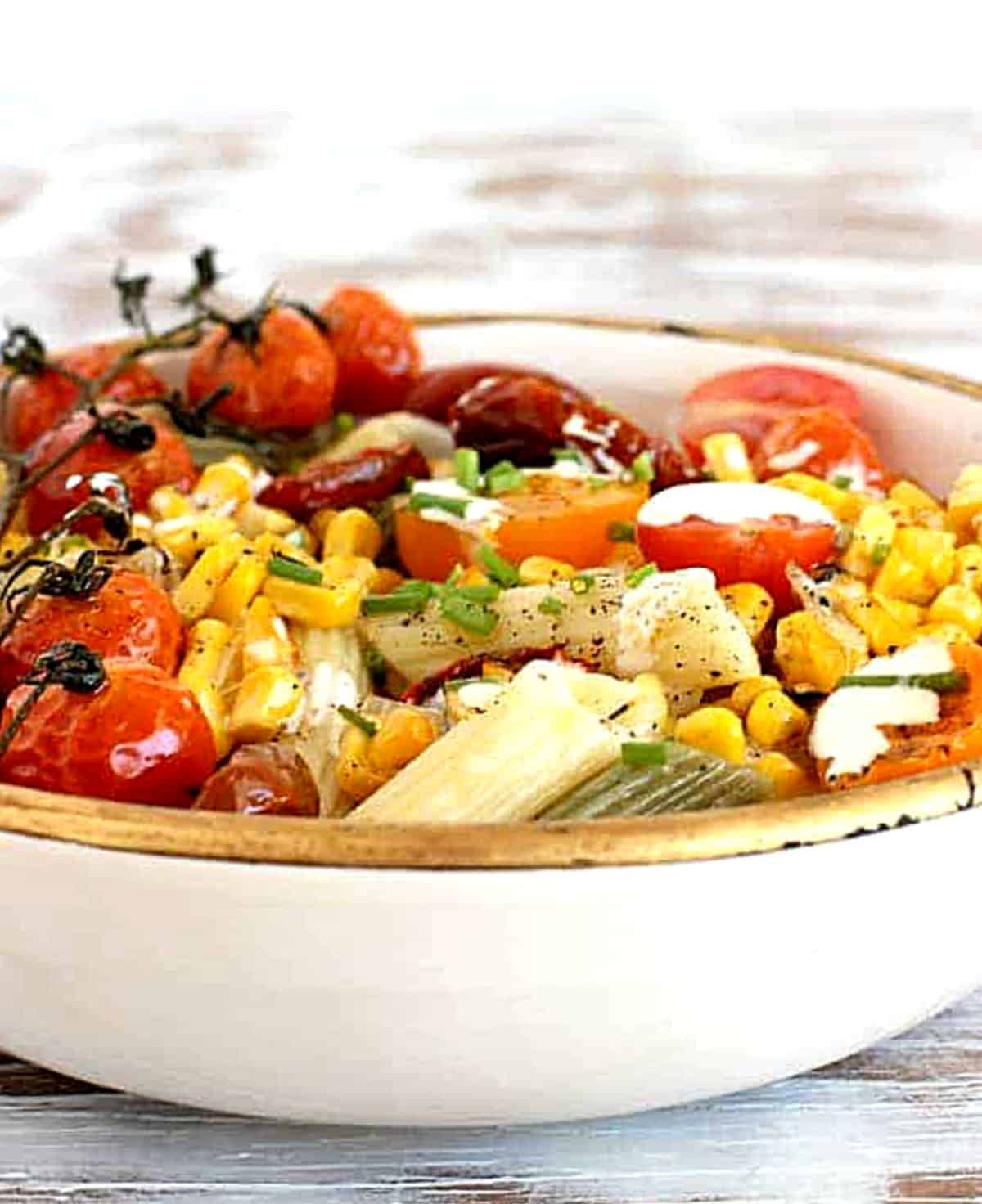 Front view of pasta plate with gold rim, mound of penne, tomatoes, corn.