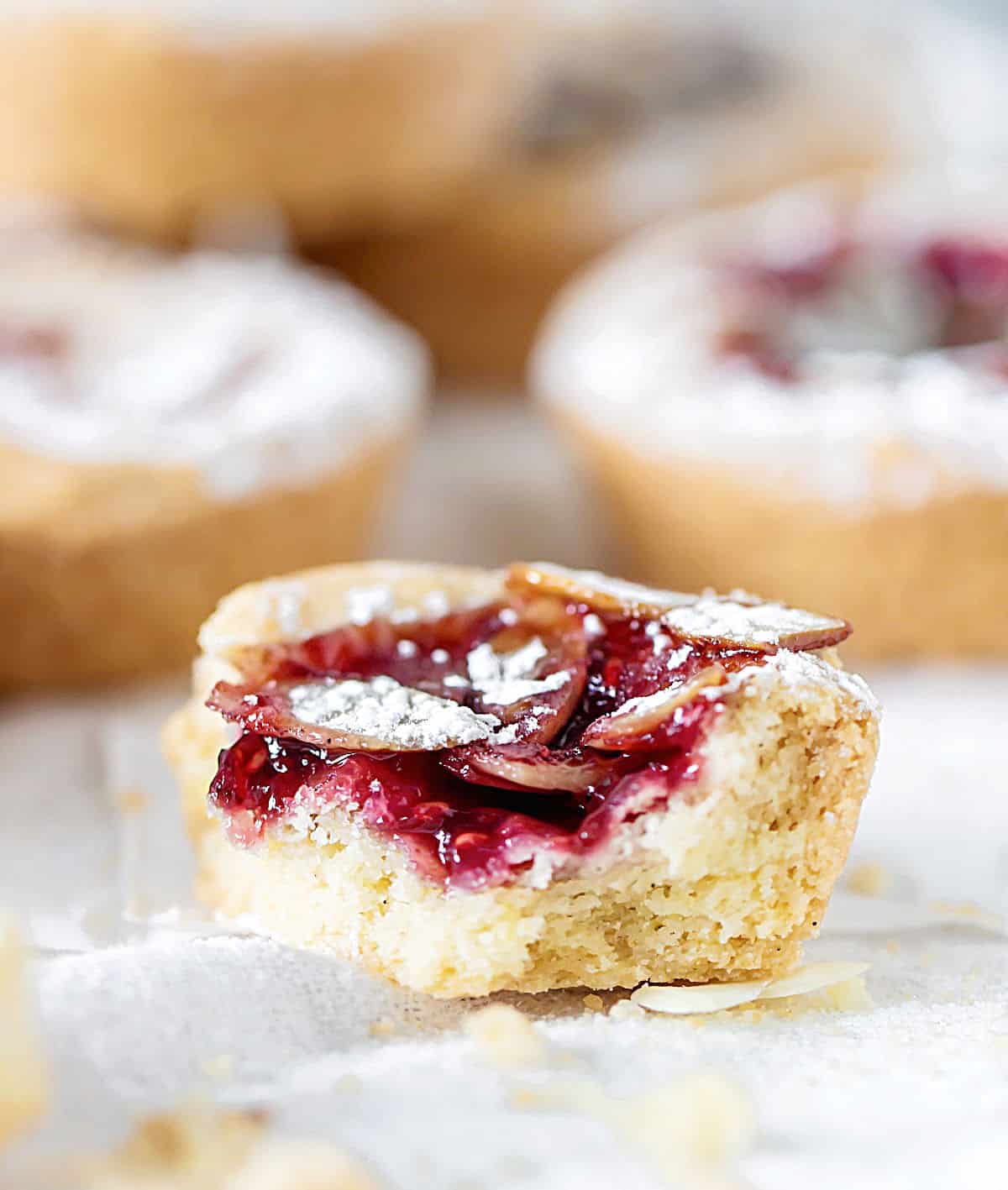 Half eaten raspberry jam tart on a white cloth with more blurred tart in the background.