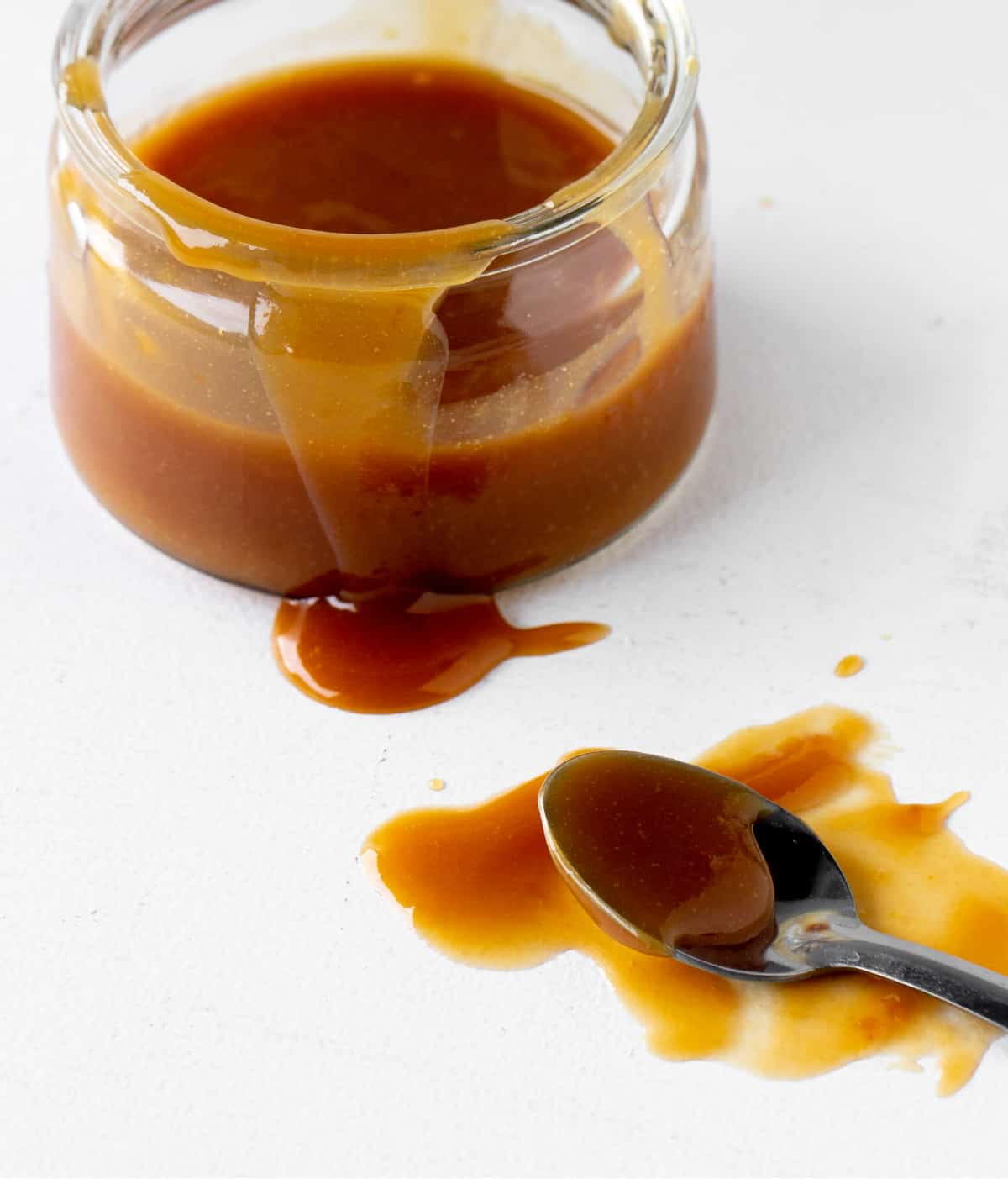 Silver spoon and glass jar dripping dulce de leche on white surface