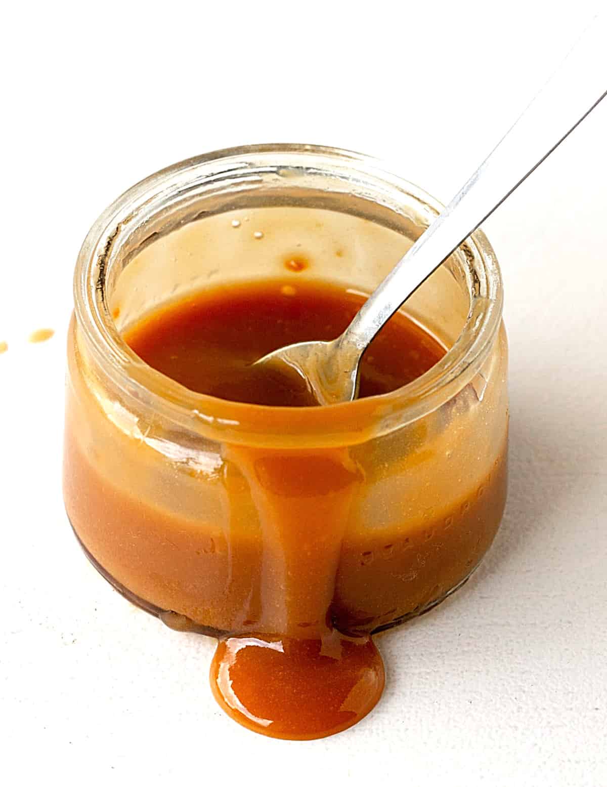 Jar of dripping dulce de leche with silver spoon inside; white surface