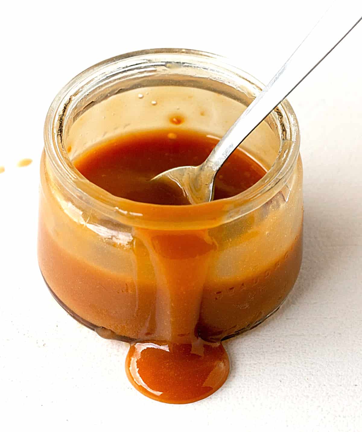 Glass jar with dripping caramel sauce, spoon inside, white background