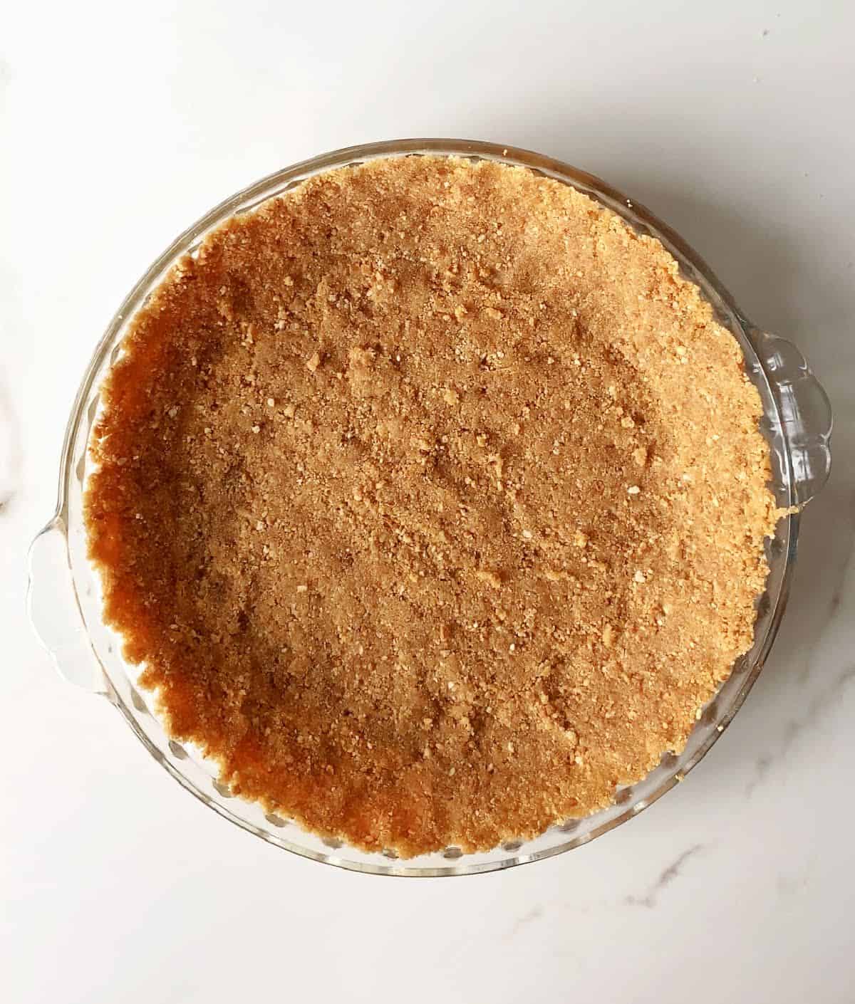 Graham cracker crust in a glass pie dish on a white marble surface. Top view.