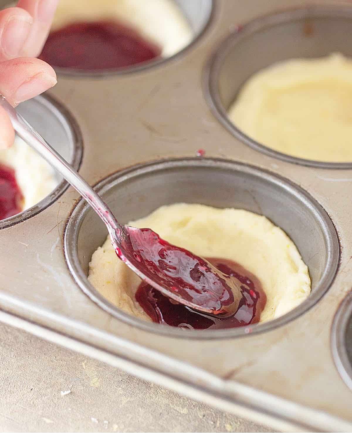 Filling cookie dough cups in a muffin pan with raspberry jam. 