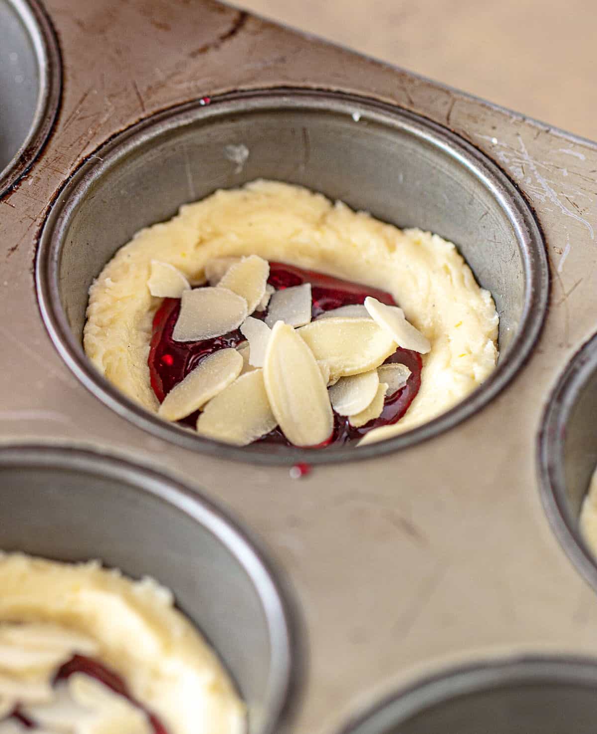 Almond topped jam filled unbaked cookie cups in metal muffin pan.