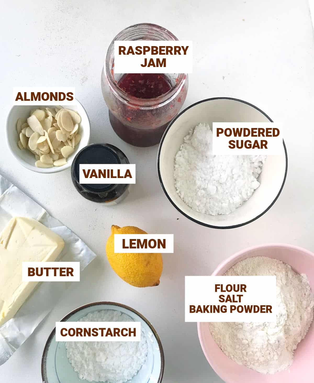 On a white surface bowls with raspberry jam cookies ingredients including a lemon, almonds, butter, powdered sugar, flour, cornstarch.