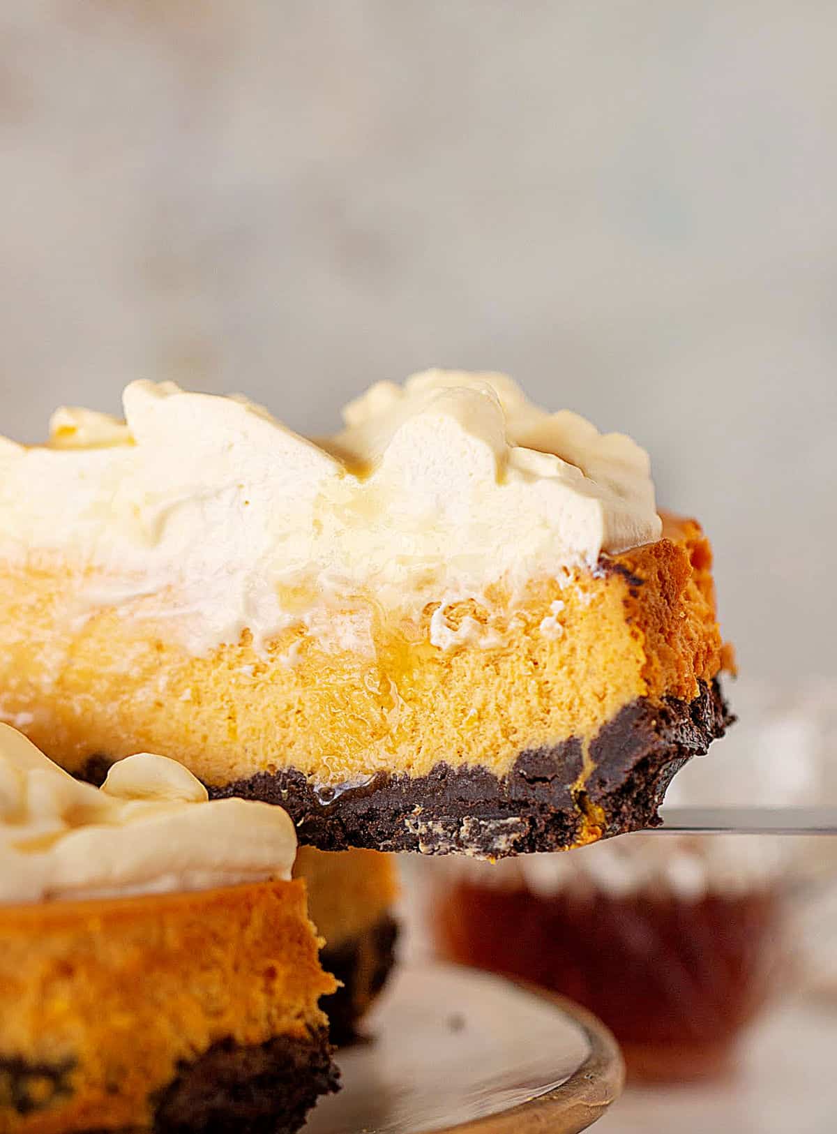 Slice of pumpkin cheesecake with whipped cream and chocolate crust being lifted from whole cake. Grey background.