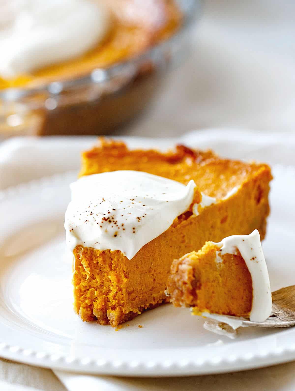 A single slice of pumpkin pie with cream on top on a white plate, a fork with a bit beside it.