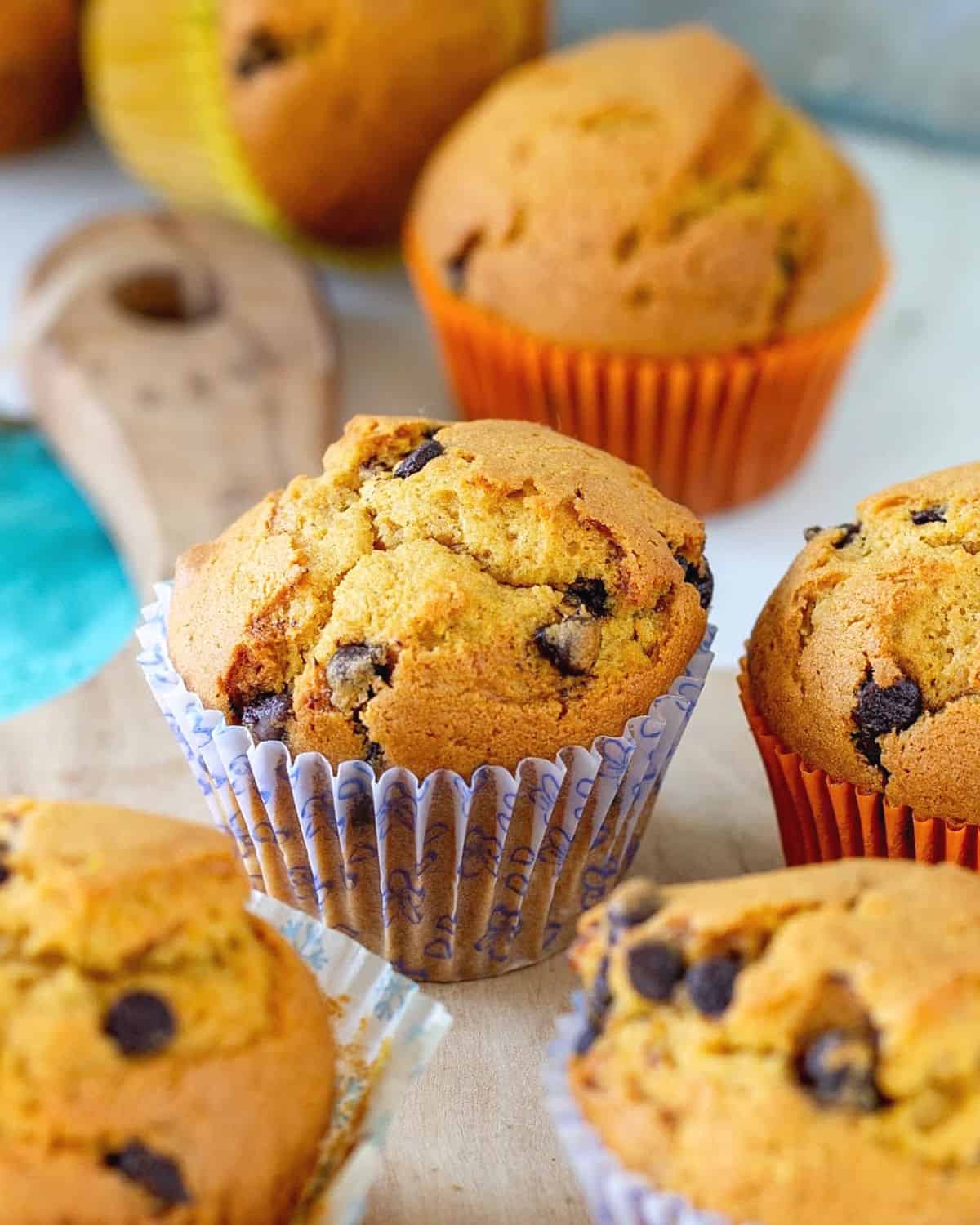 Several chocolate chip pumpkin muffins on a light wooden board, colorful paper cups