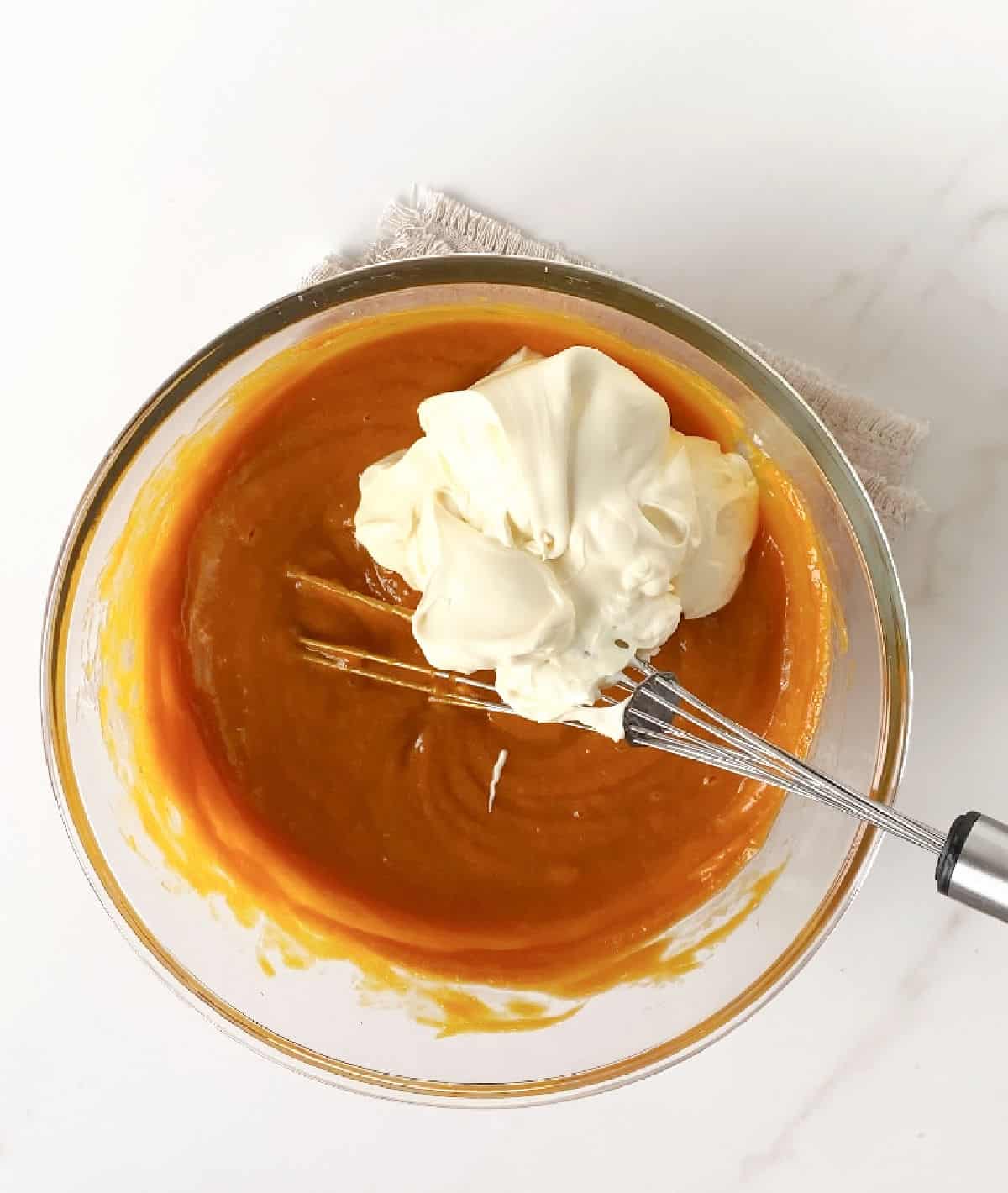 Sour cream and cream added to pumpkin pie filling in a glass bowl with a wire whisk. White marble surface.