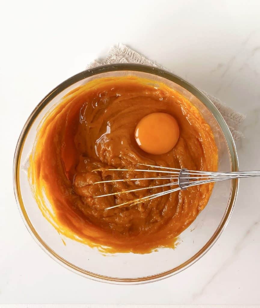 Egg added to pumpkin pie filling in a glass bowl with a whisk. White marble surface.