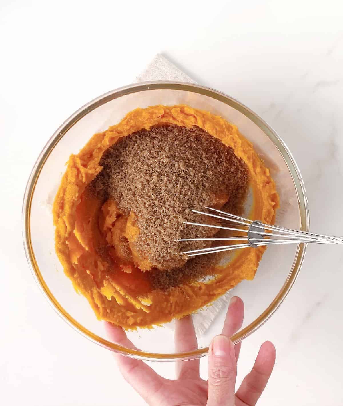 Brown sugar added to pumpkin puree in a glass bowl with a whisk. Hand holding it. White surface. 