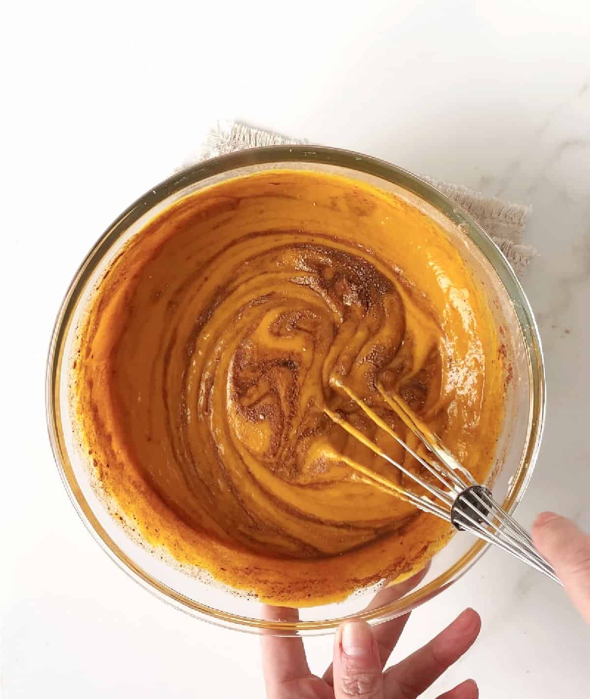 Top view of pumpkin pie filling with spices being whisked in a glass bowl on a white surface.
