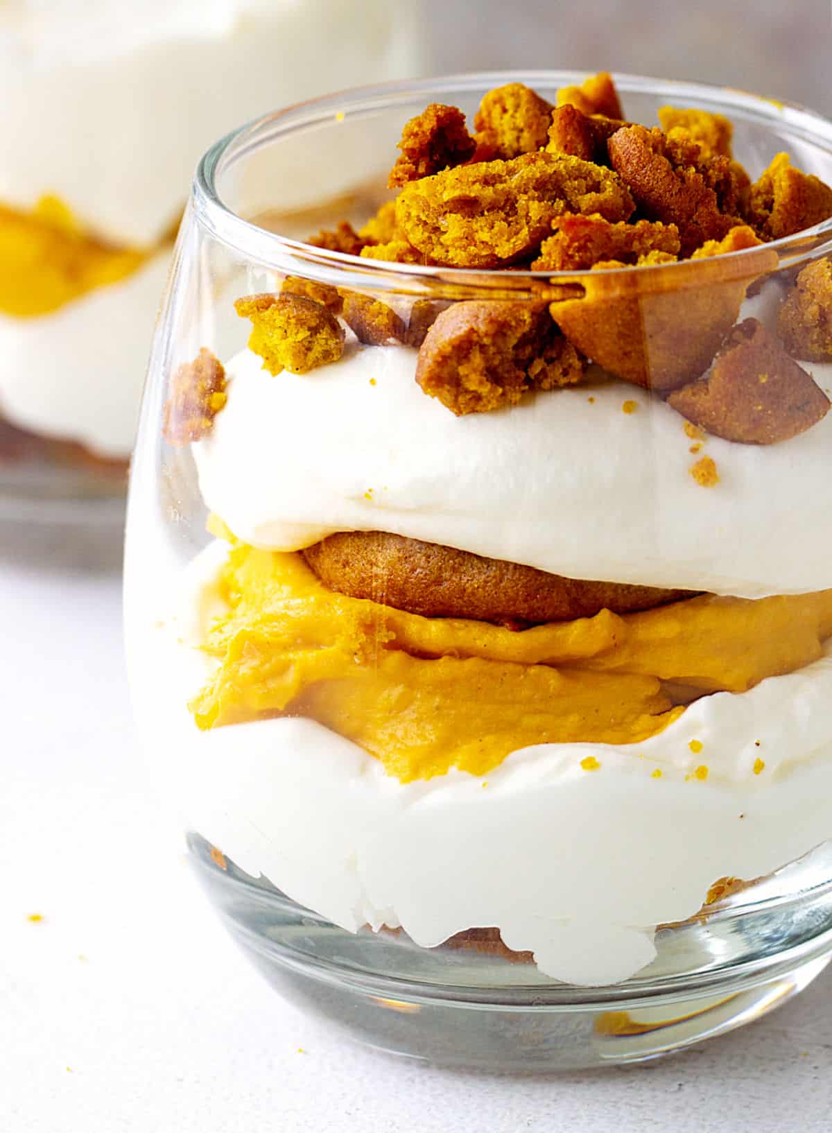 Close-up of glass with pumpkin cake, whipped cream, and pumpkin cheesecake; white surface.