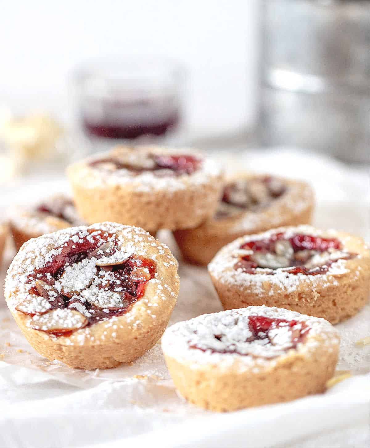 Pile of jam tarts with powdered sugar on a white cloth. Metal sifter and glass in the background.