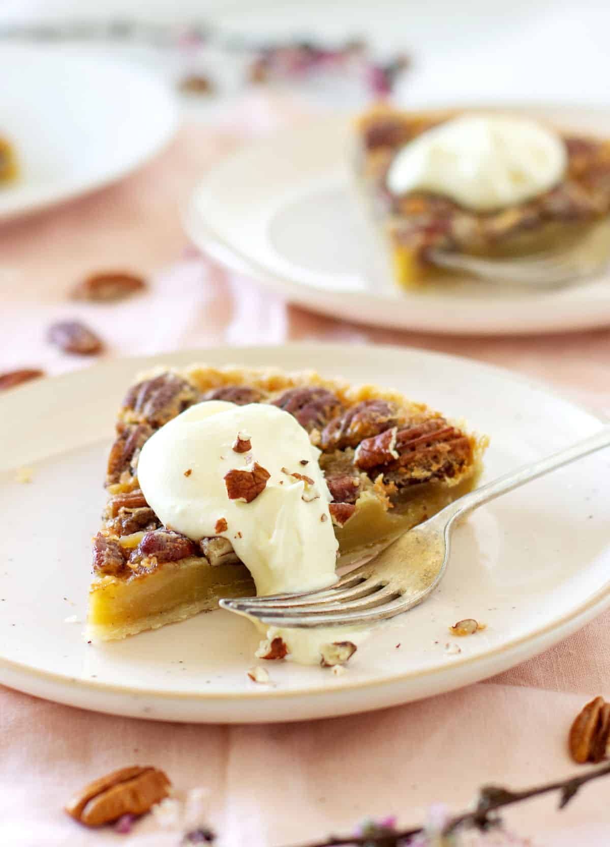 Slices of pecan pie with whipped cream on pink plates and embroidered tablecloth.