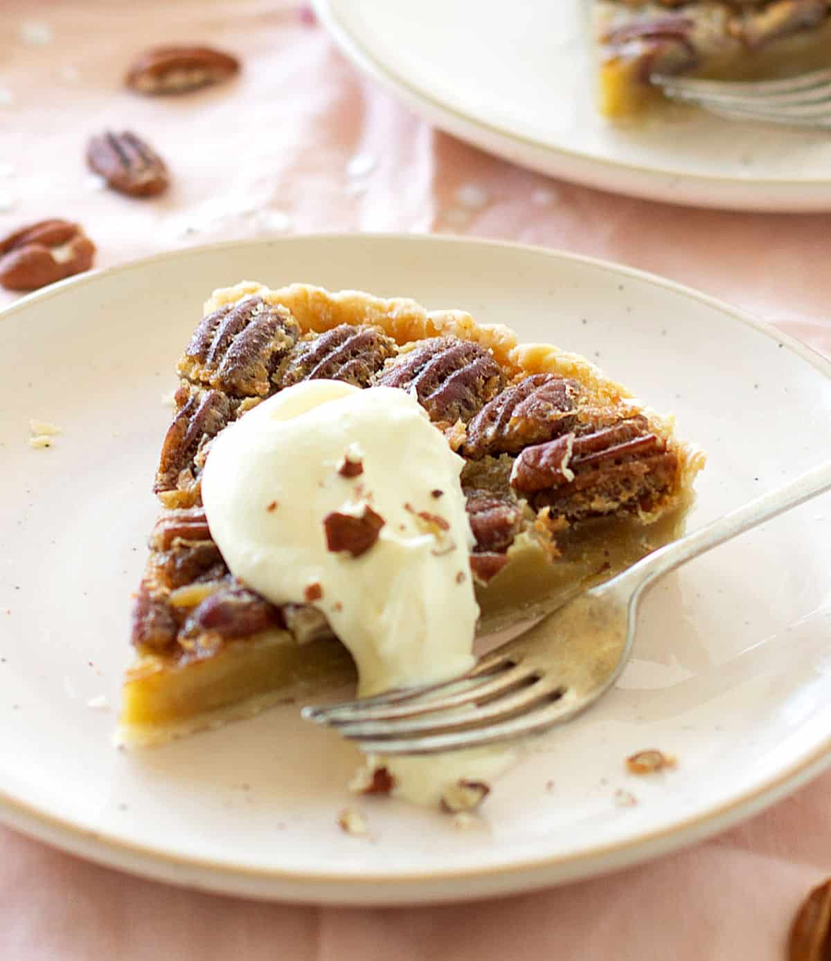 Single slice of pecan pie in light pink plate, dollop of cream, fork. Pink tablecloth.