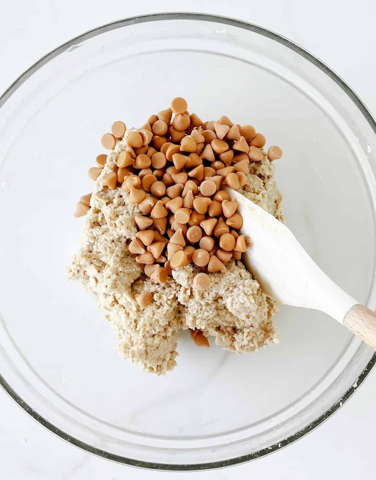 Butterscotch chips added to oatmeal cookie dough in a glass bowl with white spatula.