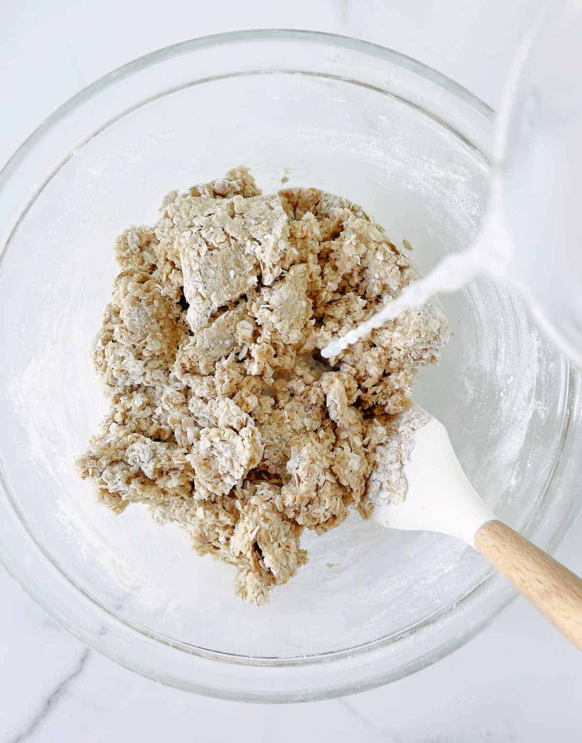 Milk being poured over oatmeal cookie mixture in a glass bowl. A white wooden spatula.