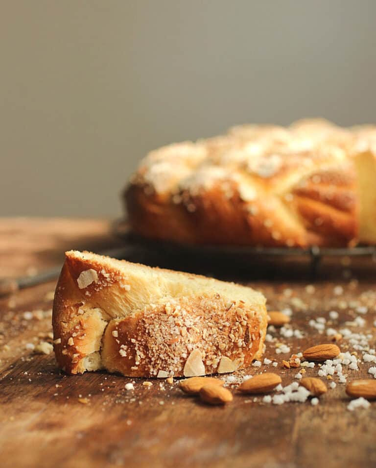 On a wooden table a slice of sweet bread with crunchy topping, scattered almonds, whole braid in background