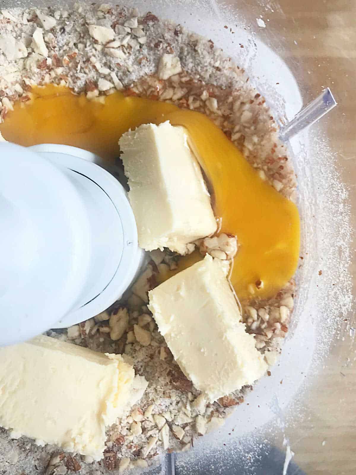 Butter pieces and egg added to almond mixture in a food processor's bowl. View from above. 