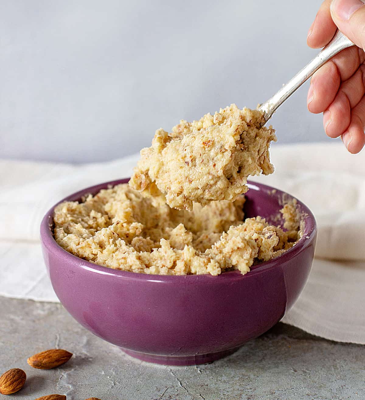 Hand lifting spoon with almond cream from purple bowl, grey background, loose almonds.