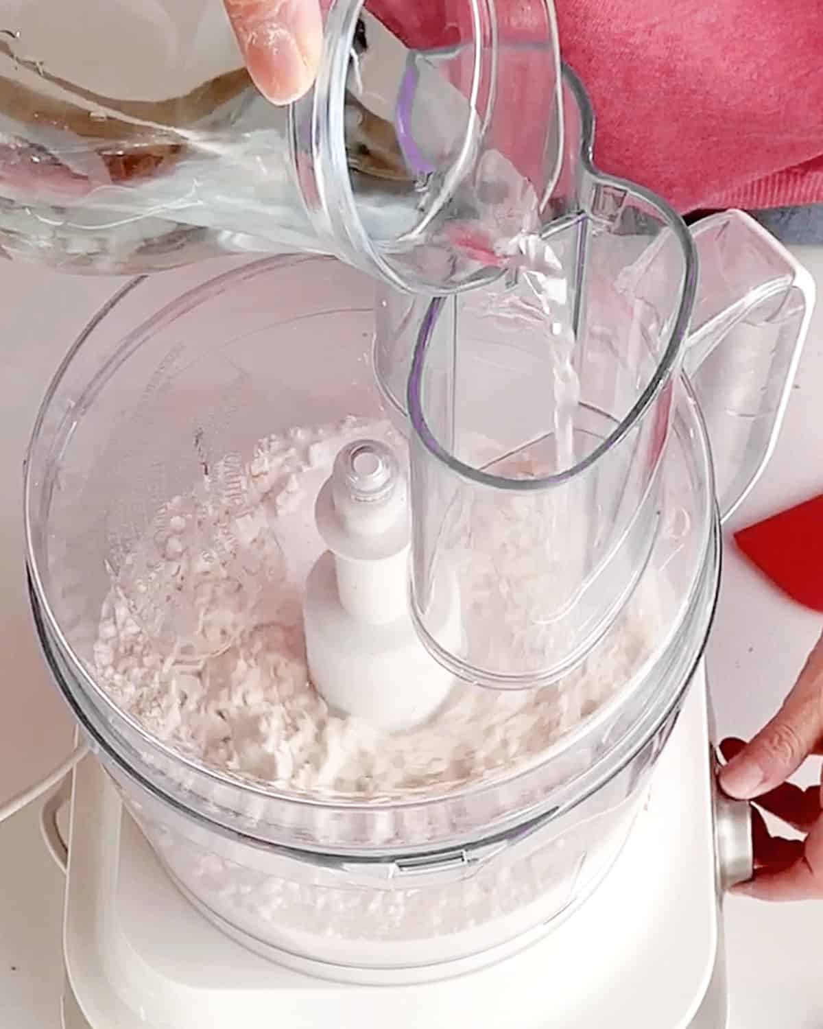 Adding water through feed tube of a food processor to flour mixture.