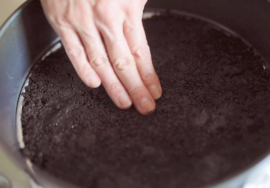 Patting an oreo crust mixture on a round cake pan.