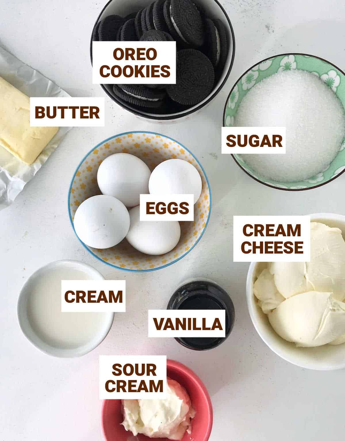 White surface with colorful bowl containing Oreo cheesecake ingredients such as eggs, cream cheese and butter.