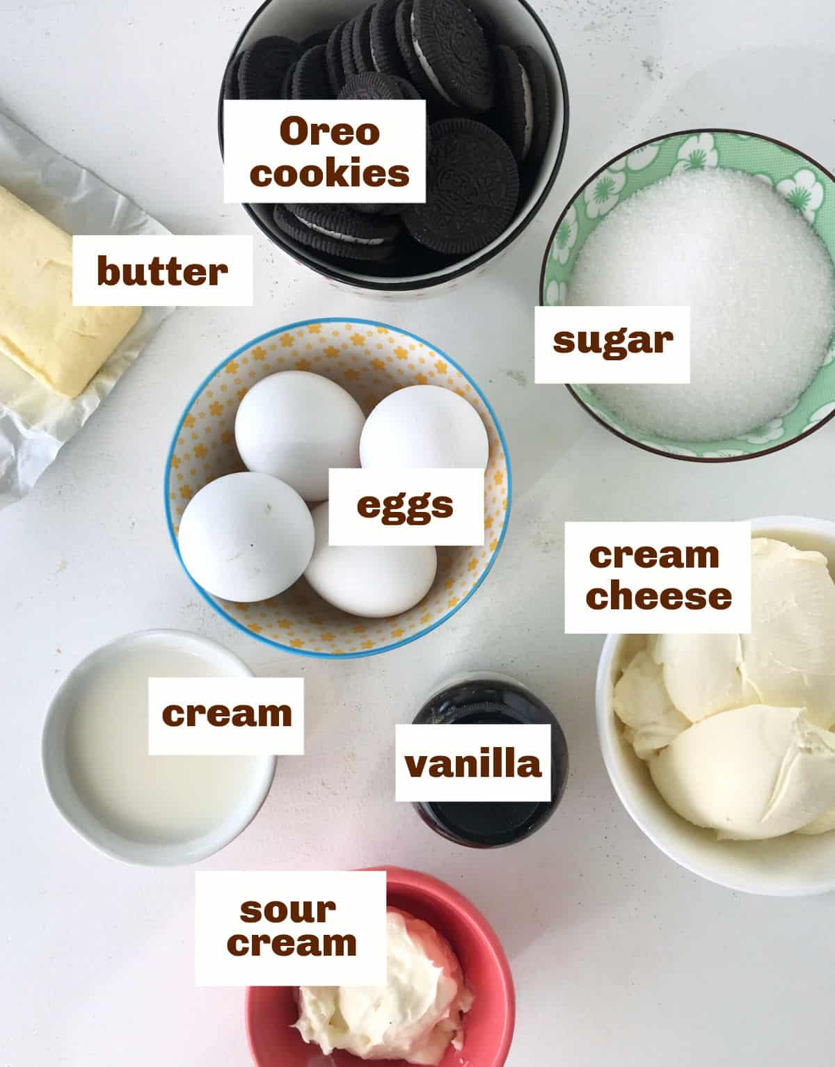 White surface with colorful bowl containing Oreo cheesecake ingredients such as eggs, cream cheese and butter