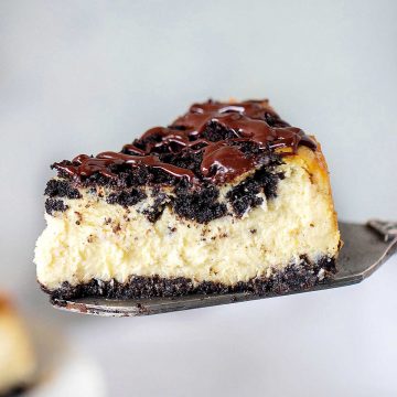Slice of ganache topped oreo cheesecake lifted with a metal cake server. Grey background.