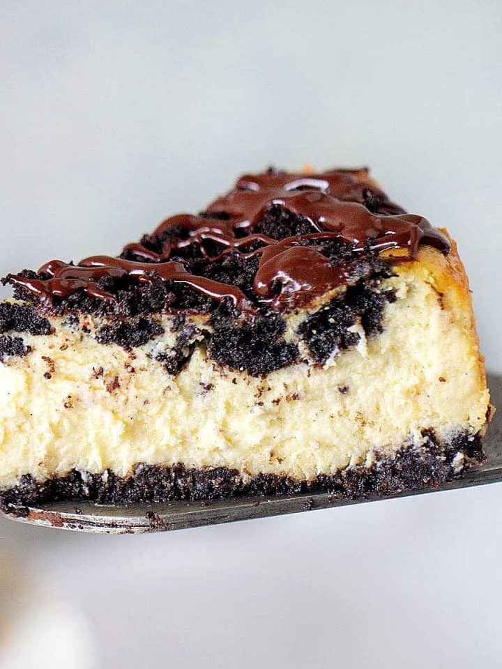 Slice of ganache topped oreo cheesecake lifted with a metal cake server. Grey background.