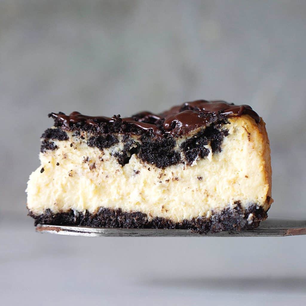 One slice of chocolate topped oreo cheesecake on a metal cake server, grey background