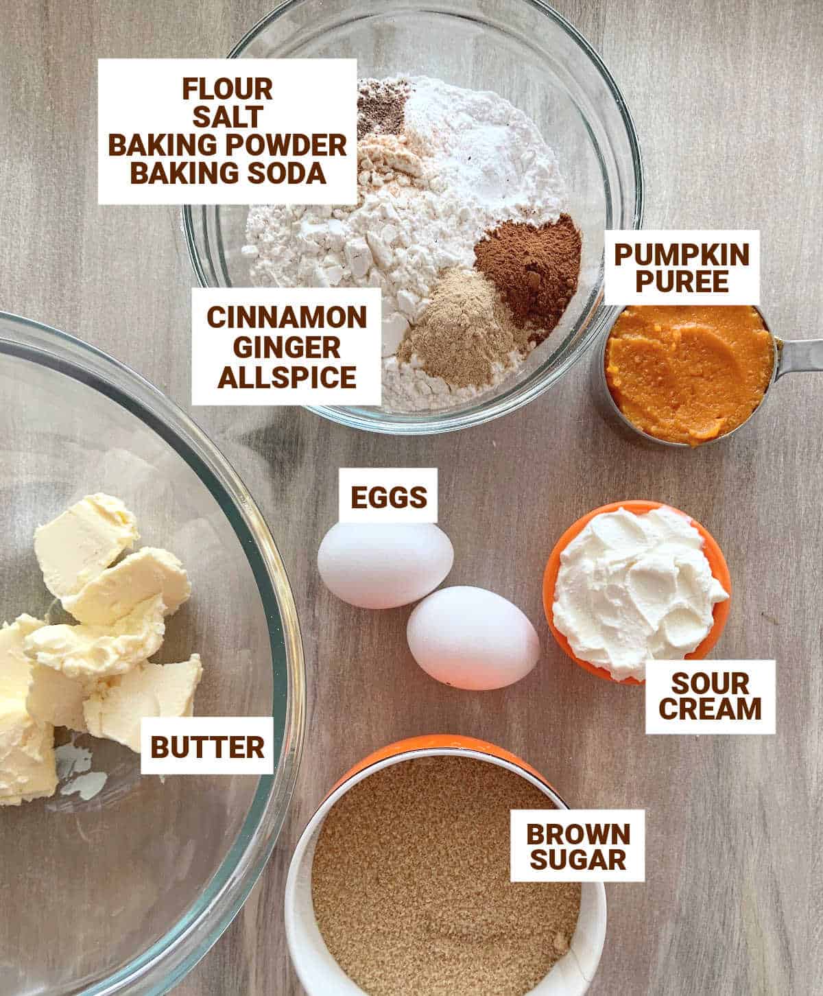 Brownish surface with bowls containing pumpkin cake ingredients, including eggs, sour cream, spices, butter, sugar, flour.
