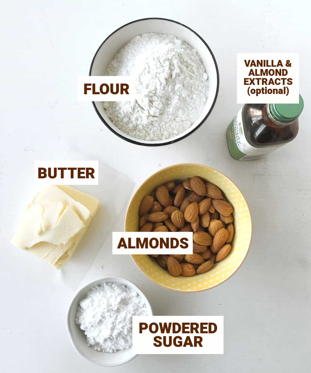 Bowls with almonds, flour, sugar, chunk of butter and a small bottle on a white surface.