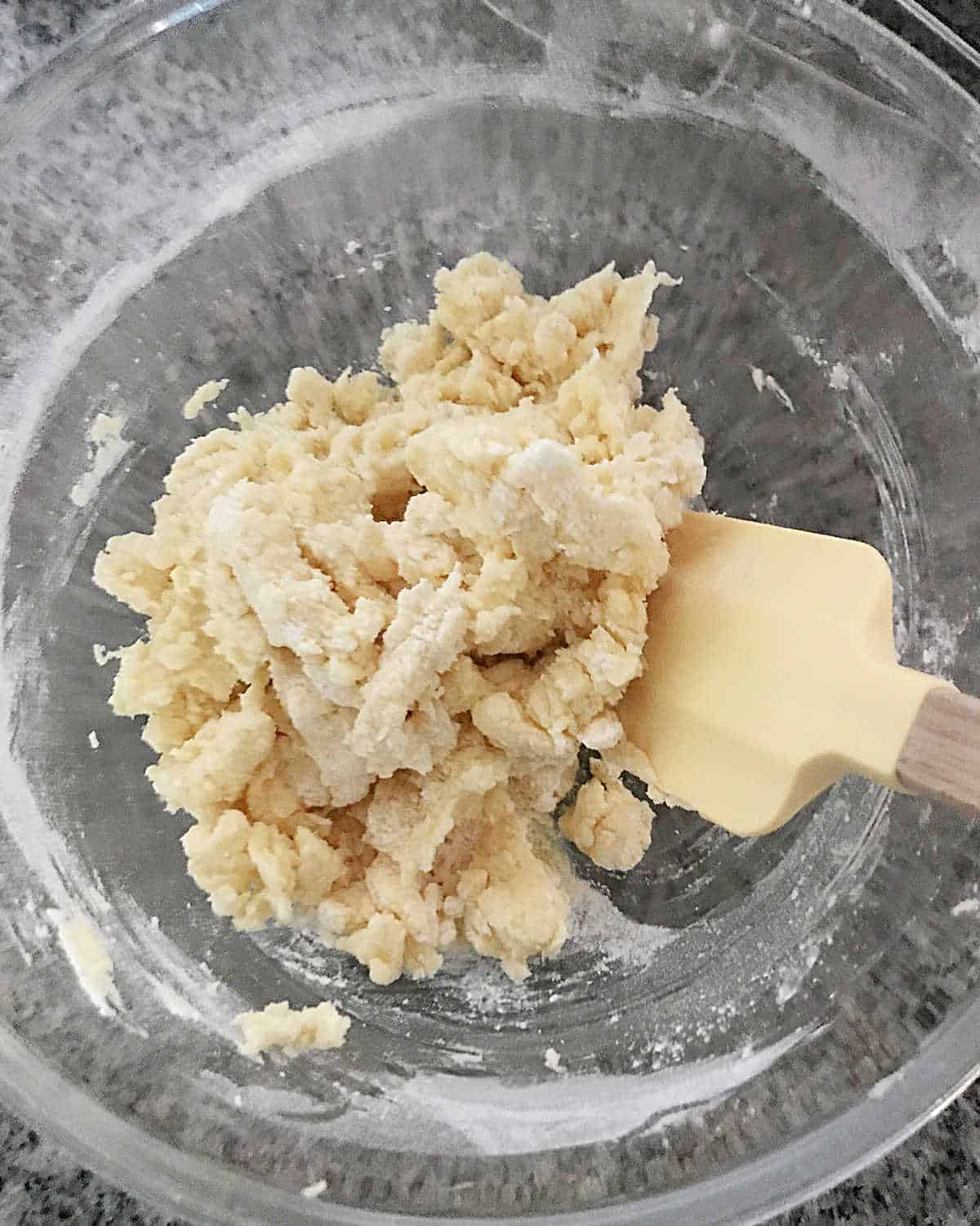 Mixing cookie dough in a glass bowl with a yellow spatula. Grey surface.