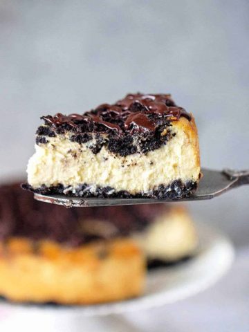 Metal cake server with slice of Oreo cheesecake. Rest of cake blurred against the grey background.