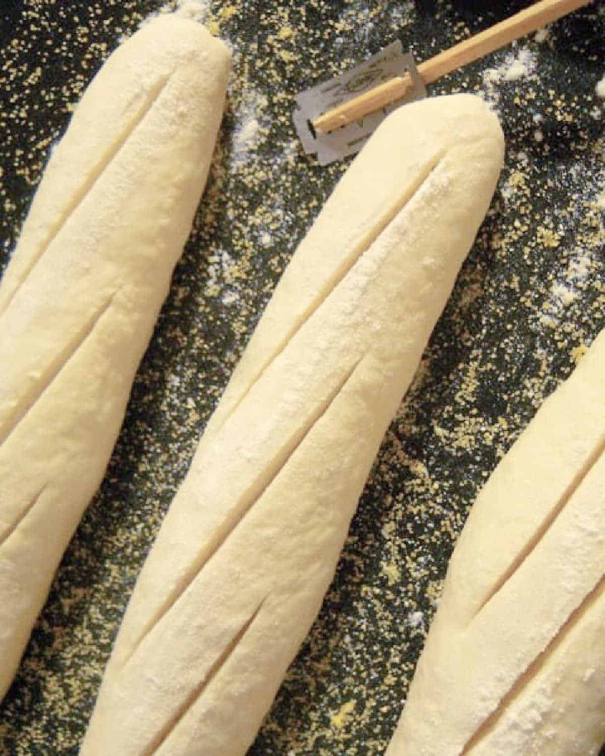 Scored baguettes on a dark baking pan with a lame (razor) beside them.