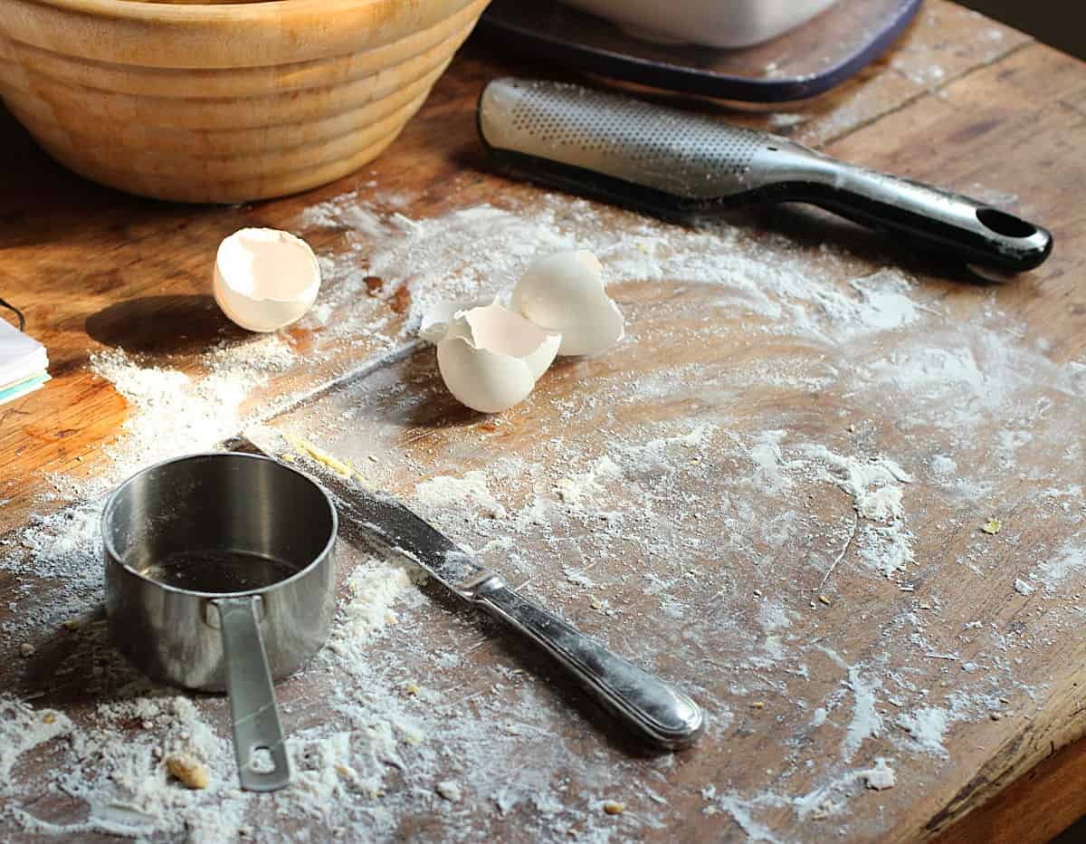 Wood table with flour, egg shells, knife, metal cup and grater