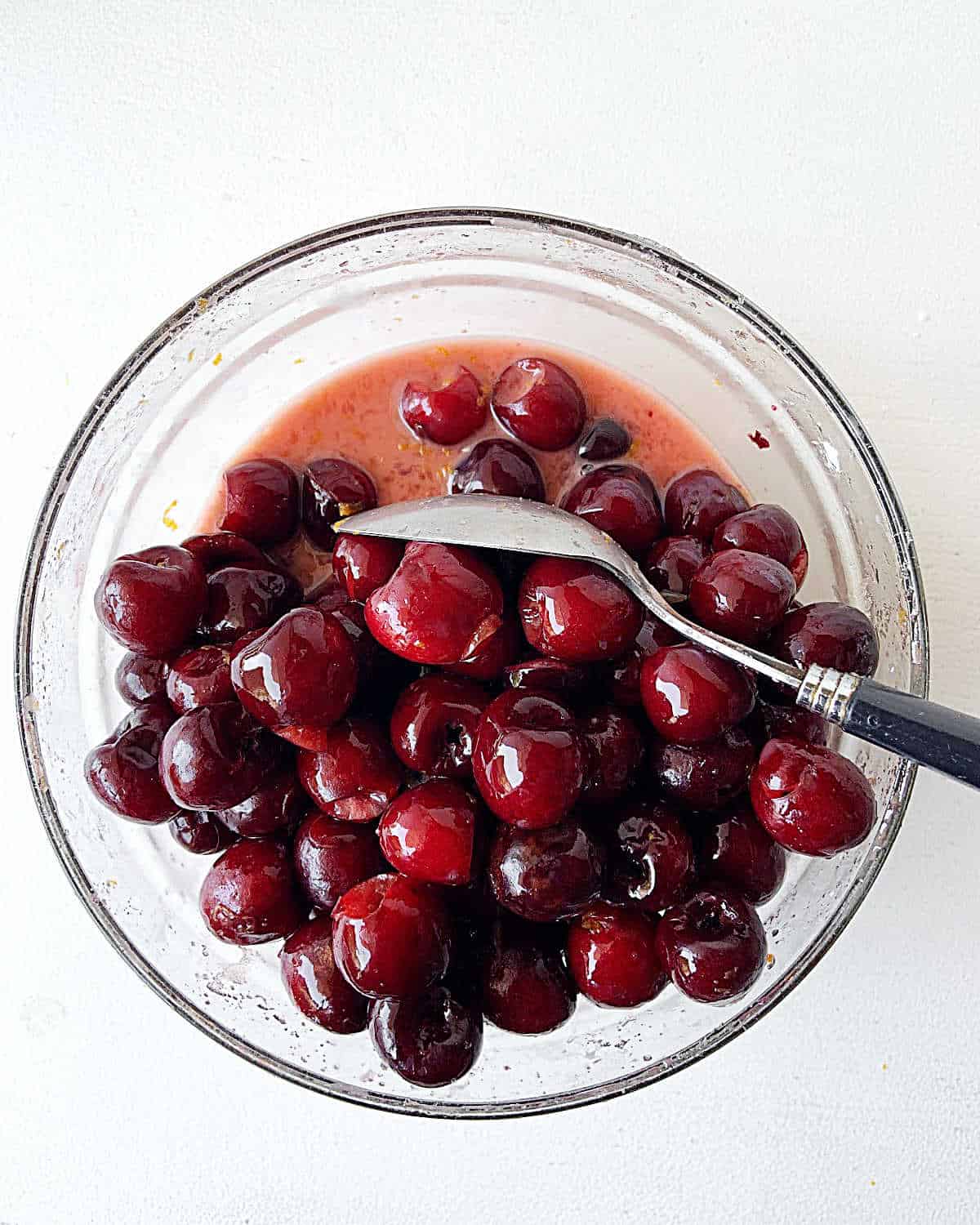 Juicy cherry filling in a glass bowl with a spoon on a white surface.