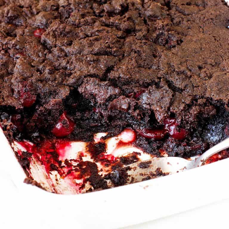 White baking dish with chocolate cherry dump cake. A serving missing, silver spoon inside.