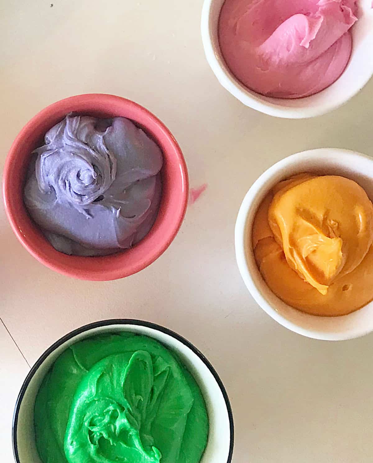 Four bowls with cream cheese frosting in different colors, white surface
