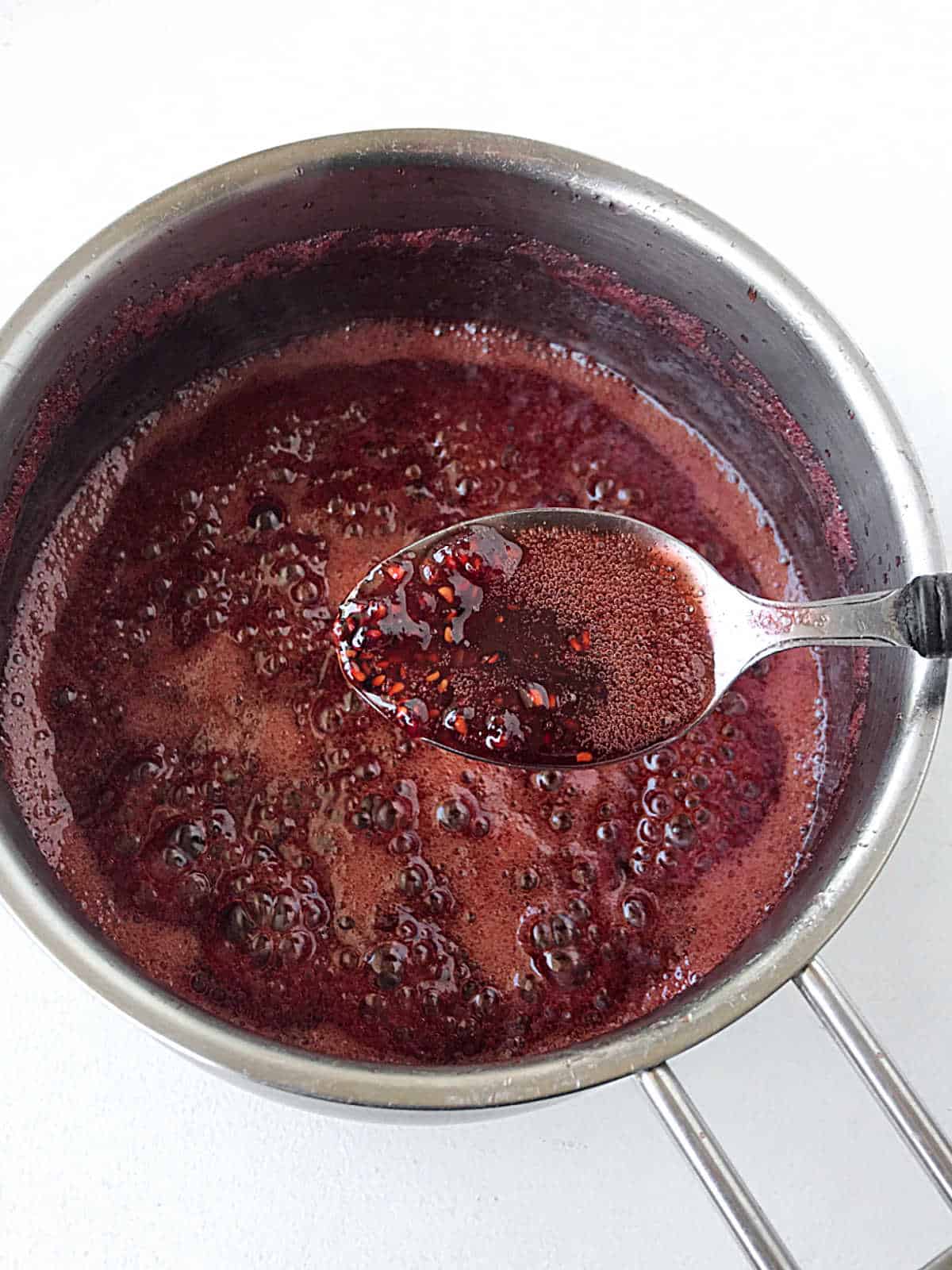 Cooked raspberry filling in a spoon with metal saucepan below with more filling. White surface.