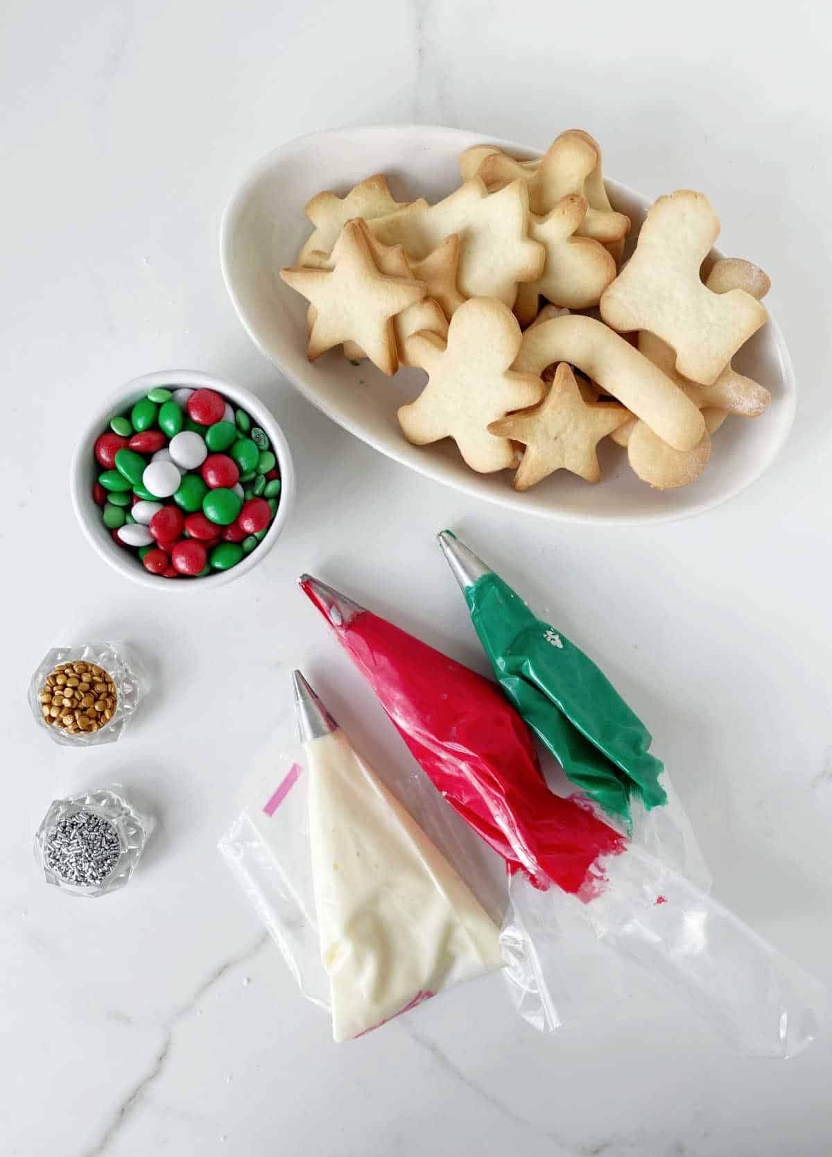 Piping bags with Christmas colored frosting, bowl with sugar cookies, sprinkles. White marbled surface.