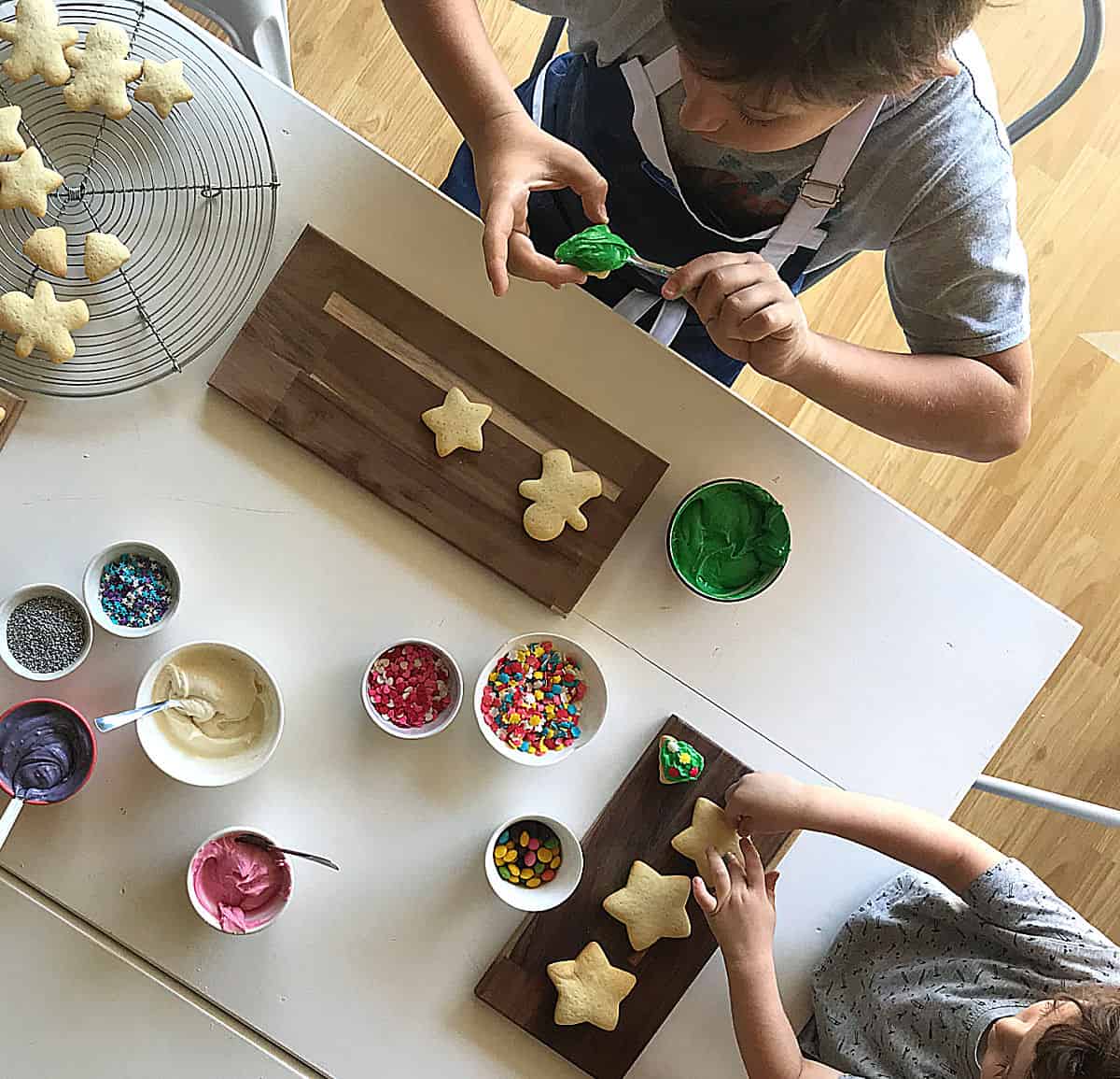 Top view of kids decorating sugar cookies on white table, bowls with colored frosting and sprinkles