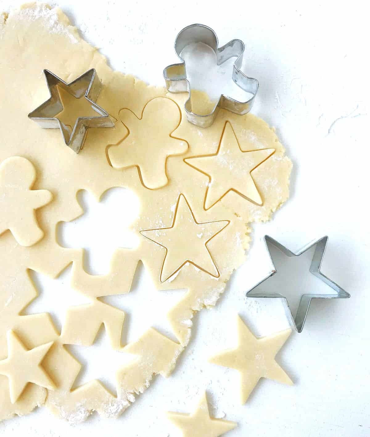 Rolled sugar cookie dough with cut outs and metal cookie shapes, on white surface.